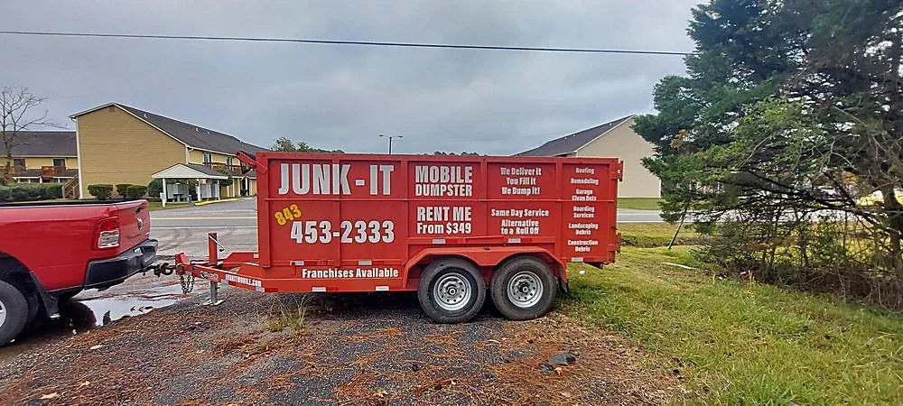 A Junk It mobile dumpster set level on grass beside a building, resting on its rubber tires