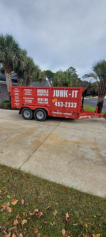 A Junk It mobile dumpster on a residential driveway with palmettos behind it